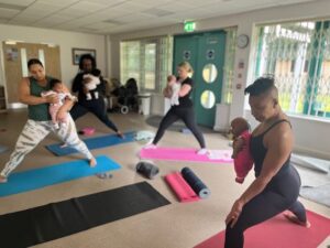 Three women standing doing yoga with their babies led by an instructor at the front of the room.