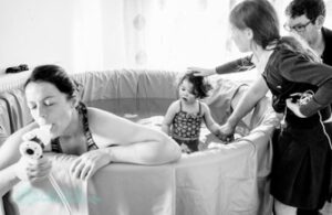 Black and white photo of mum in labour in birthing pool using gas and air. Toddler is stood in pool and dad and older daughter stood at the side.