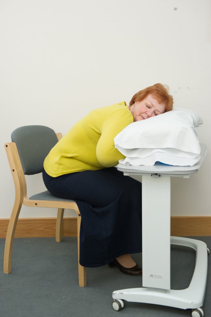 Photograph of a person sitting in a chair, leaning forward with their head reasting on a table with a pillow on it