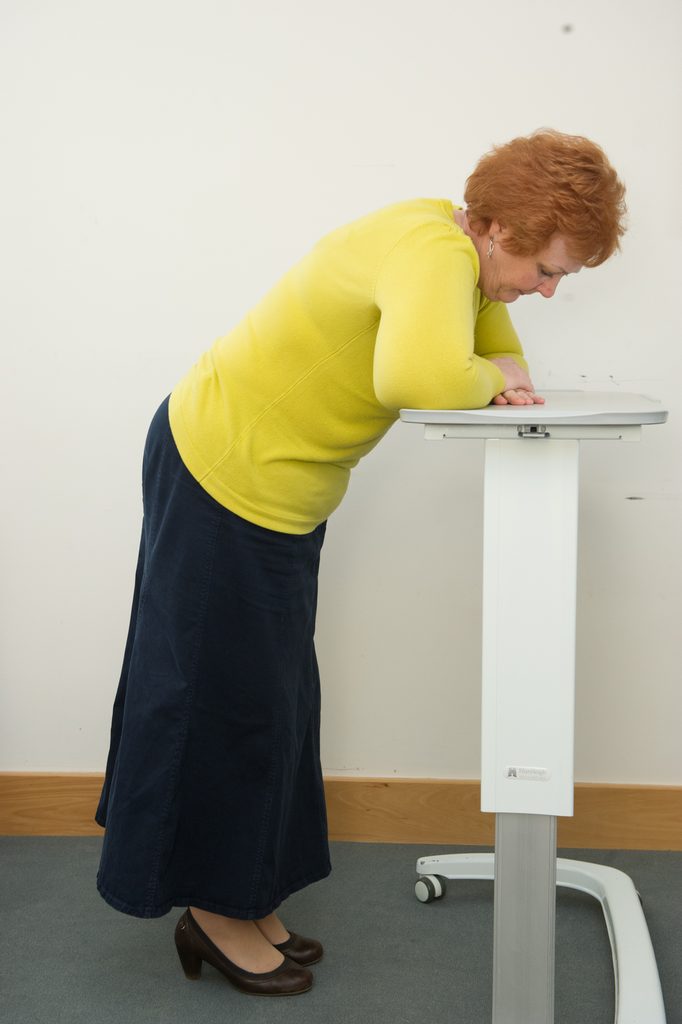 Photograph of a person stood, resting their arms on a tall table, leaning forward