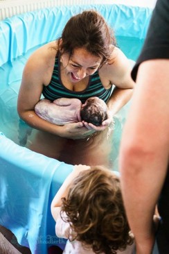 Mum holding a newborn baby in the birth pool smiling at her toddler who is stood at the side.