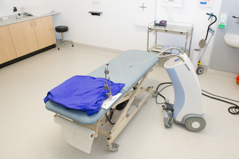 Image shows Brachytherapy Treatment Room where you will have your treatment. 

Contains the brachytherapy treatment machine next to the treatment couch.

This is the machine which will deliver your treatment.

Before treatment the radiation source is stored safely  in a safe inside this machine. 

Next to the treatment unit is the treatment couch where you will lie to have your treatment.

On the couch is a pillow or headrest.

There is a metal clamp that slides under the headrest. This is to hold the treatment applicator in place whilst you lie in the couch.

Attached to the top of the machine is a white hollow wire (transfer tube)- this will be connected to the treatment applicator and enable the small radiation source to travel to the area for treatment.

The image also show a trolley on which will be your treatment template and accessories required for treatment.