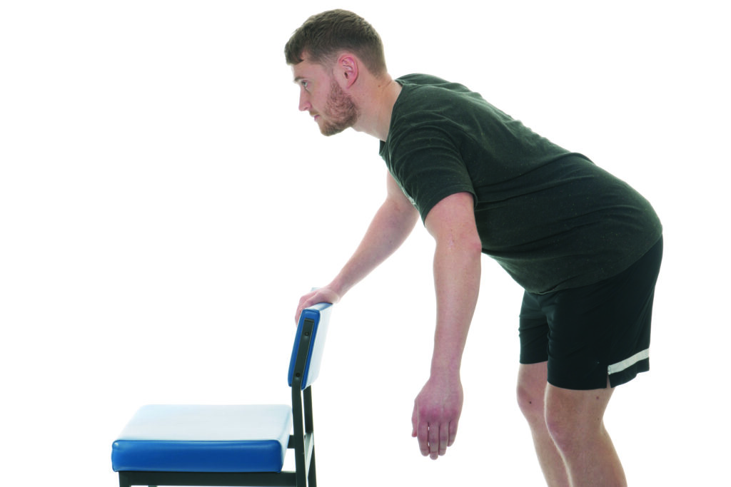 Photograph of a person leaning against a chair with one arm on the chair and one arm relaxed at side