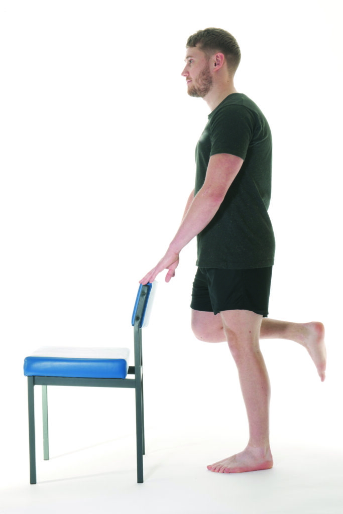 Photograph of a person stood upright holding the back of a chair with leg bent at the knee