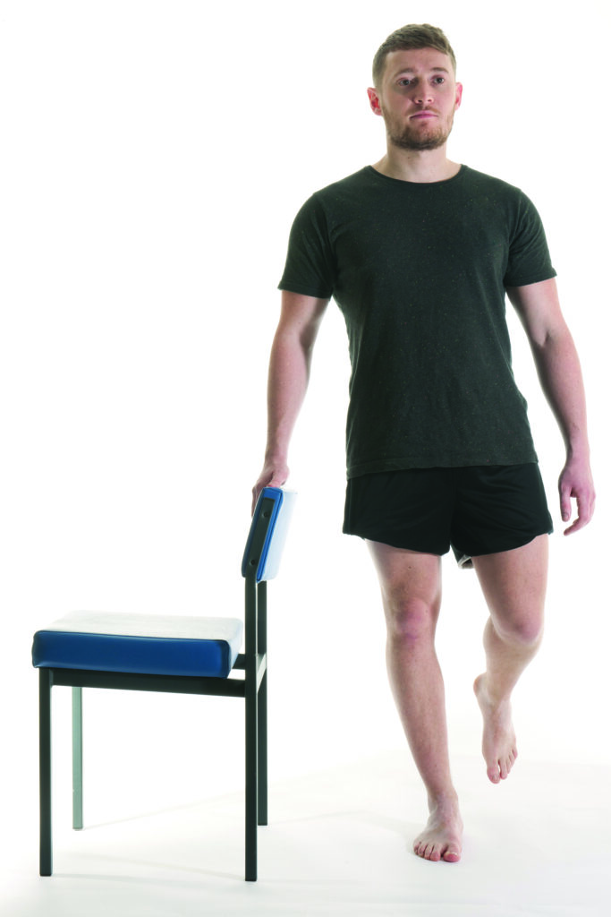 Photograph of a person stood upright holding the back of a chair side on with leg bent at the knee