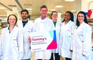 Group of 6 researchers in a laboratory wearing white lab coats. Male researcher in centre of group is holding a sign which says 'Proud to be part of the Tommy's national centre for preterm birth research.'