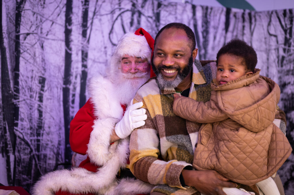 An image showing a photo been taken of Santa Claus sitting down with an adult and child both sitting on Santa Claus's lap at the Leeds Children's Hospital Christmas Lights 2023 Switch on.