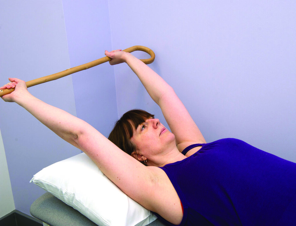 Photograph of patient with both arms up holding a walking stick.