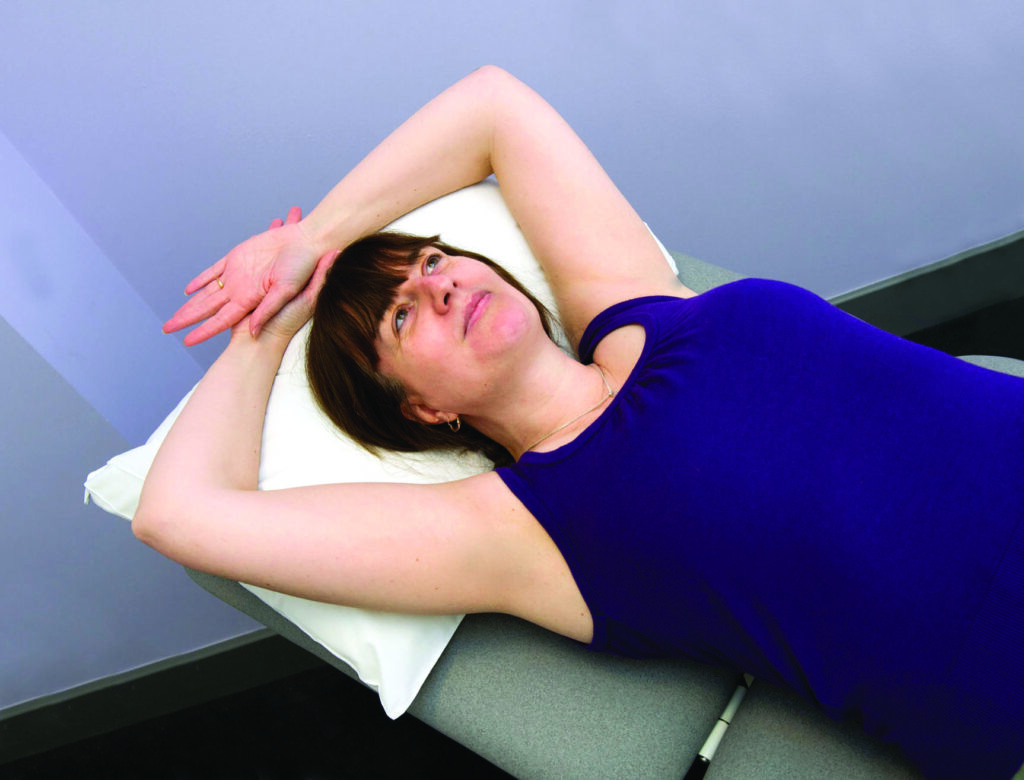Photograph of patient with both arms relaxed back onto a pillow with arms slightly bent and palms facing the ceiling.
