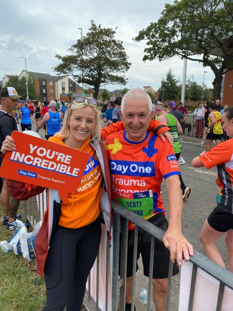 A man and a woman in bright orange Day One running gear stand at the side of a road where an official run is taking place. The woman holds  a placard saying 'You're Incredible'