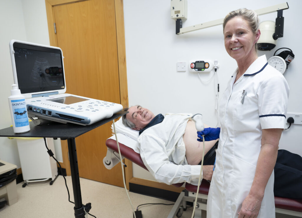 A man with his shirt open laid on a clinical couch. A woman in a white clinical uniform is stood next to him holding a scanning device with a monitor in front of her on a stand.