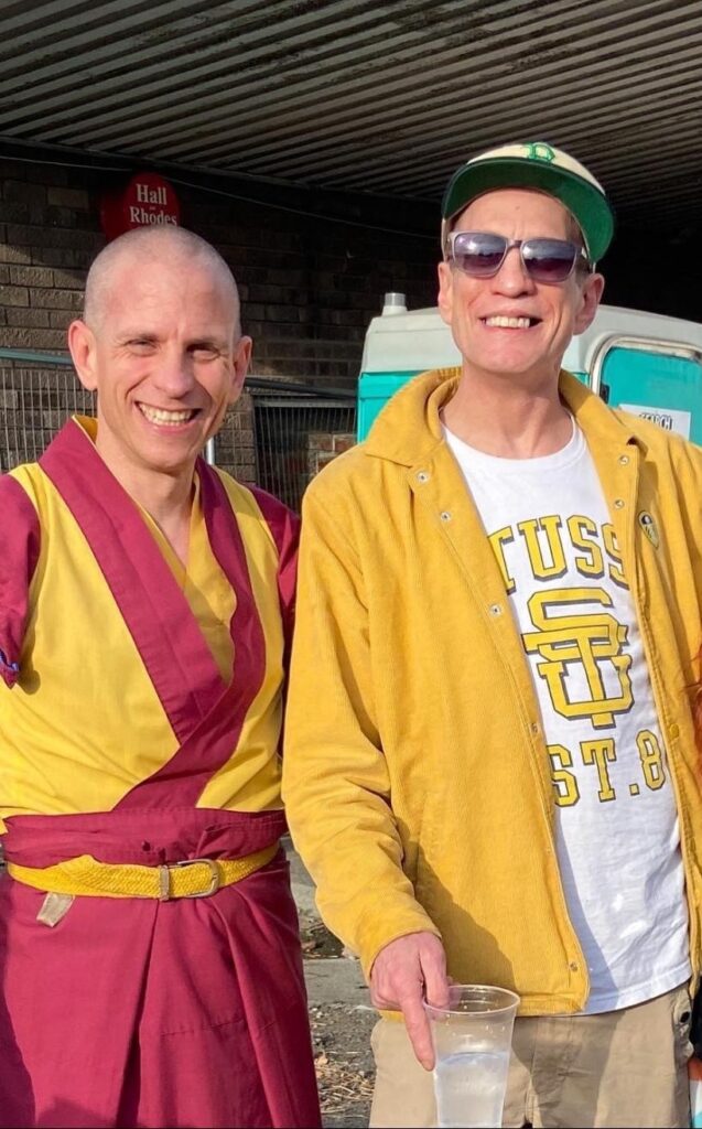 A man in yellow and burgundy robes stood next to another man in a yellow jacket, baseballl cap and sunglasses
