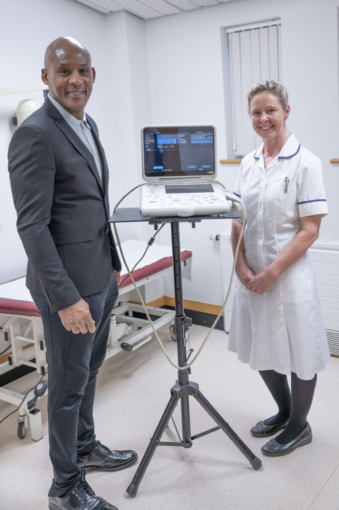 A man in a a dark grey suit stood with a woman in a white clinical uniform either side of a portable ultrasound scanner
