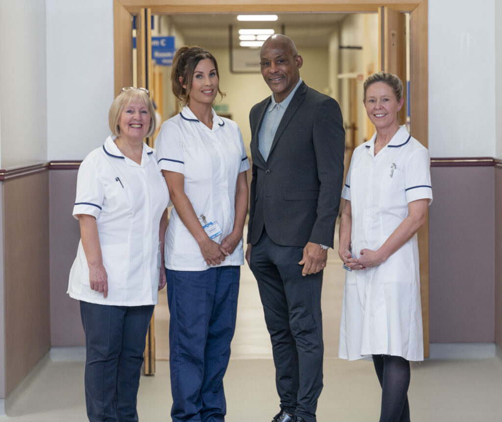 Three women in white clinical uniforms stood beside a man in a dark grey suit in a hospital corridor