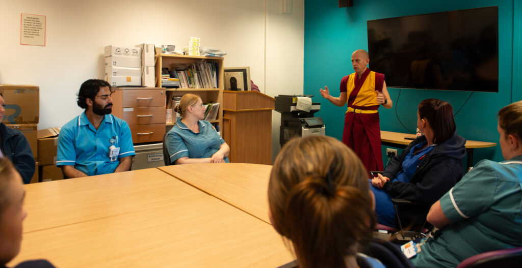 Several people in blue clinical uniforms sat around a table in an office while a man in yellow and burgundy robes speaks to them
