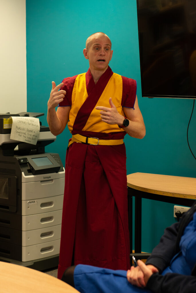 A man in yellow and burgundy robes stood in an office in front of a teal wall