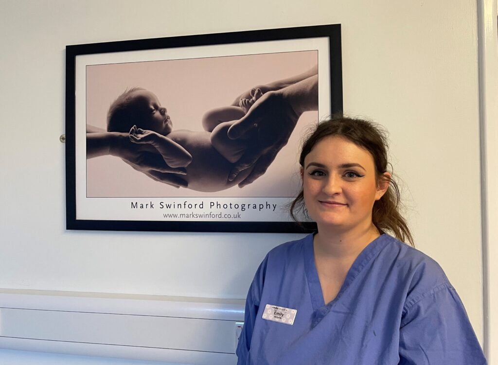 Photo of midwife in a blue top stood in front of a phot with a baby been held.