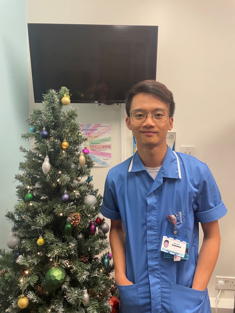 Photo of a nurser wearing glasses in a blue top next to a christmas tree.