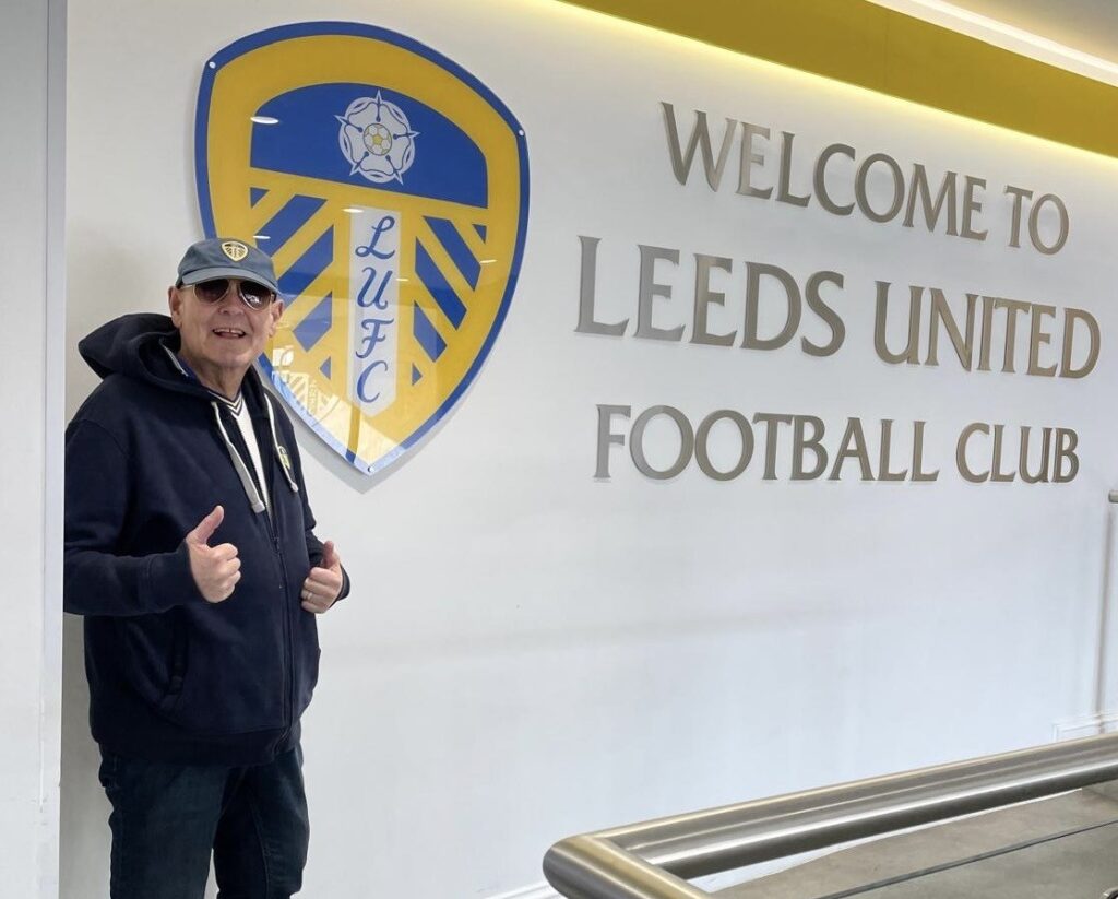 A photo of a person smiling and giving two thumbs up to the camera stood next to the a mural of the Leeds United Football logo and a sign that says "Welcome To Leeds United Football Club".