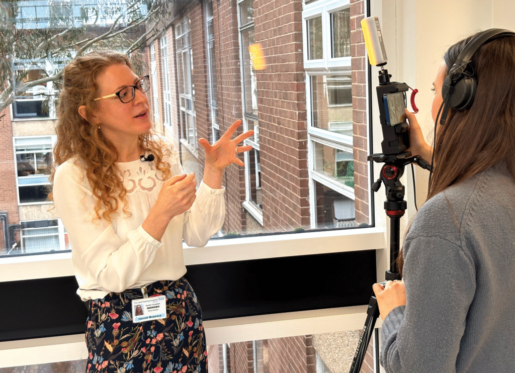 Woman wearing glasses talking to another woman who is operating a camera on a tripod. Both are standing in front of a window overlooking the rest of the building