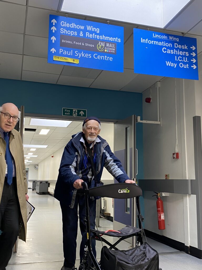 Two people in coats walking down a hospital corridor beneath blue signage. One has a mobility aid.