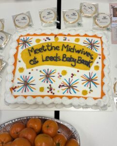 White rectangular decorated cake with 'Meet the Midwives at Leeds Baby Bank' icing decorations. Round biscuits and oranges are on the table next to the cake.