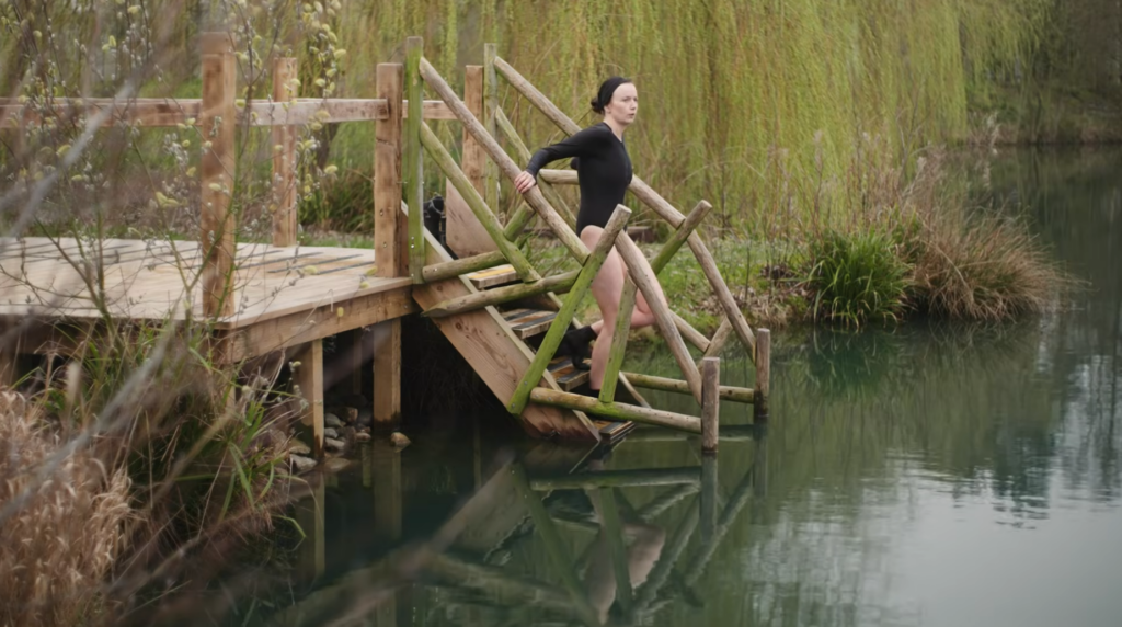 A woman in a black bathing suit stepping into a tree-fringed lake from a wooden platform