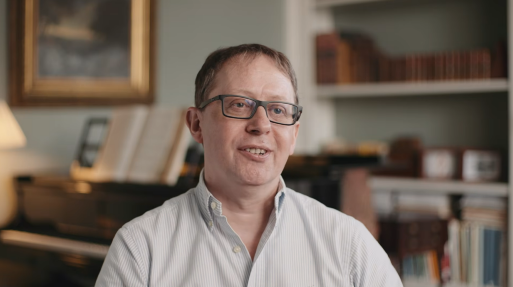 A man in a grey shirt sat in a living room with a piano and bookshelves