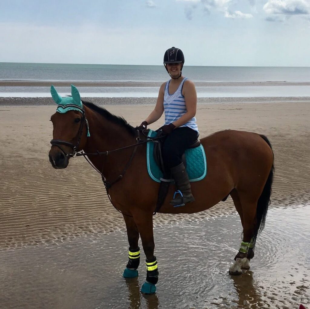 A woman in a black and white striped top and riding helmet sat upon a chestnut horse beside the sea