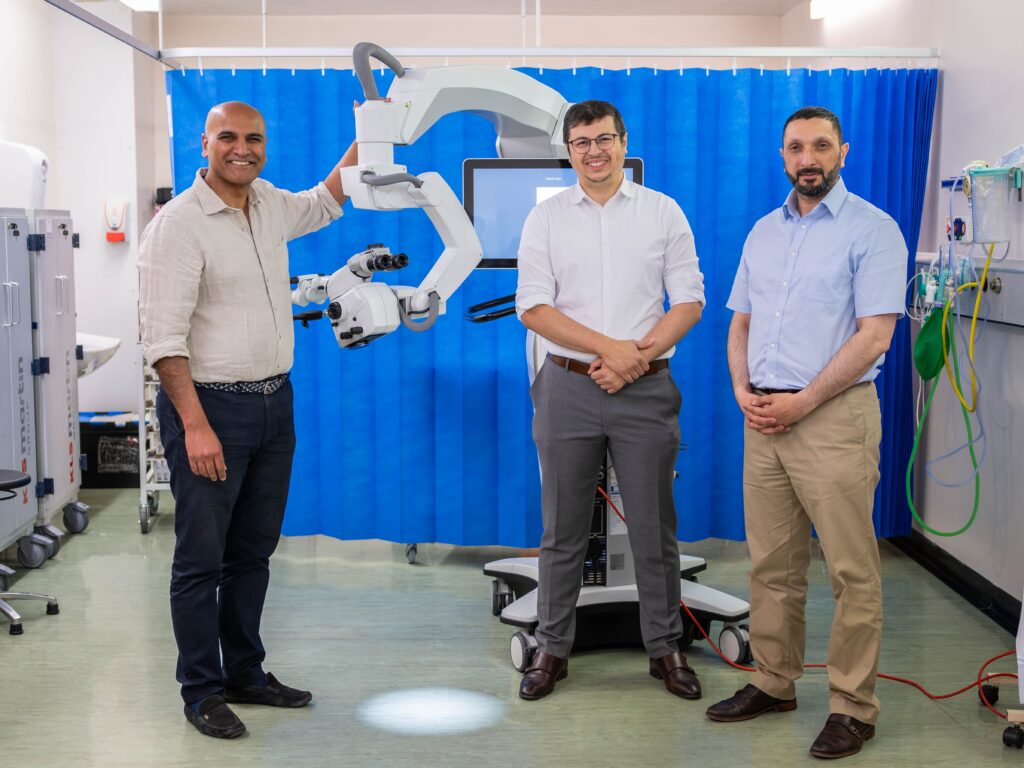 Three men in suits stood in a clinical room beside a surgical microscope