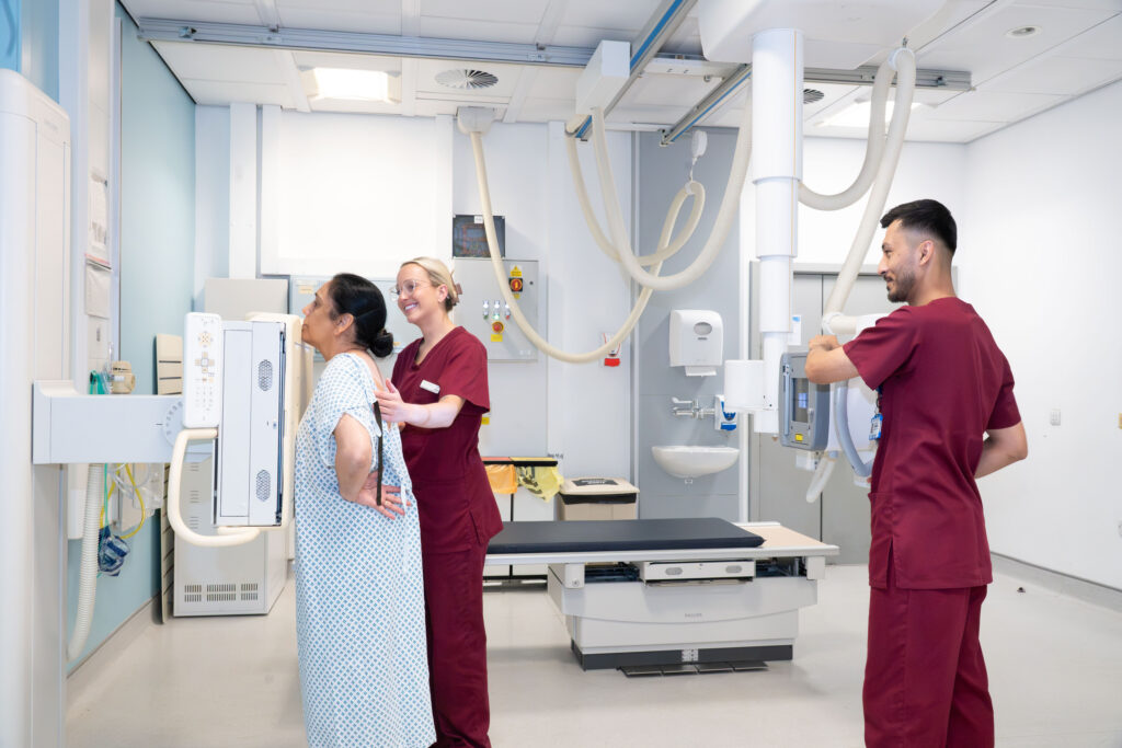 A woman in an operating gown having a chest x-ray with help from two radiographers in burgundy scrubs
