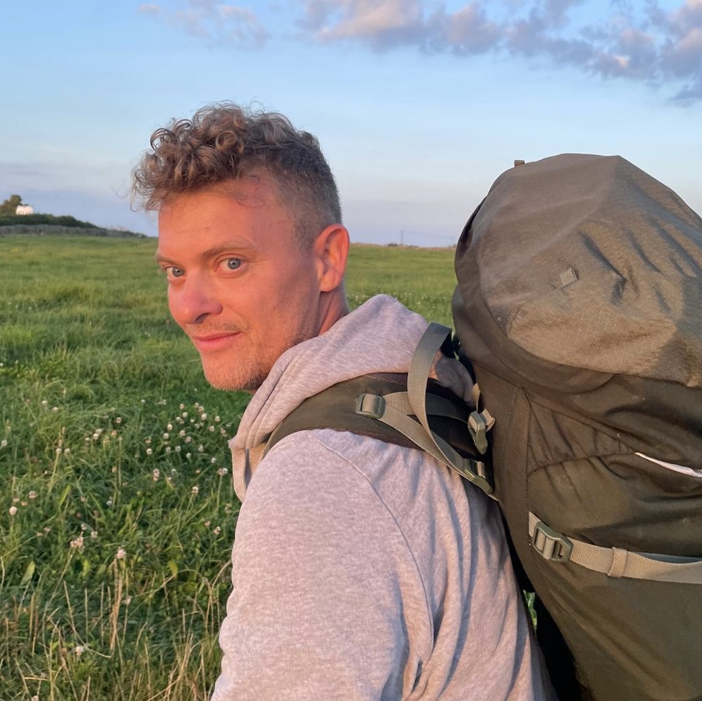 A man in a grey hoody wearing a green backpack walking through a field