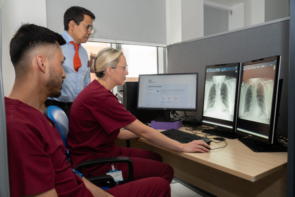 Two people in burgundy scrubs and a man in a white shirt and red tie looking at X-ray scans on a computer screen