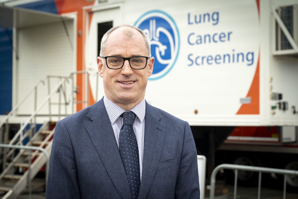 A man in a blue suit and glasses stood in front of a white trailer featuring the words 'lung cancer screening'