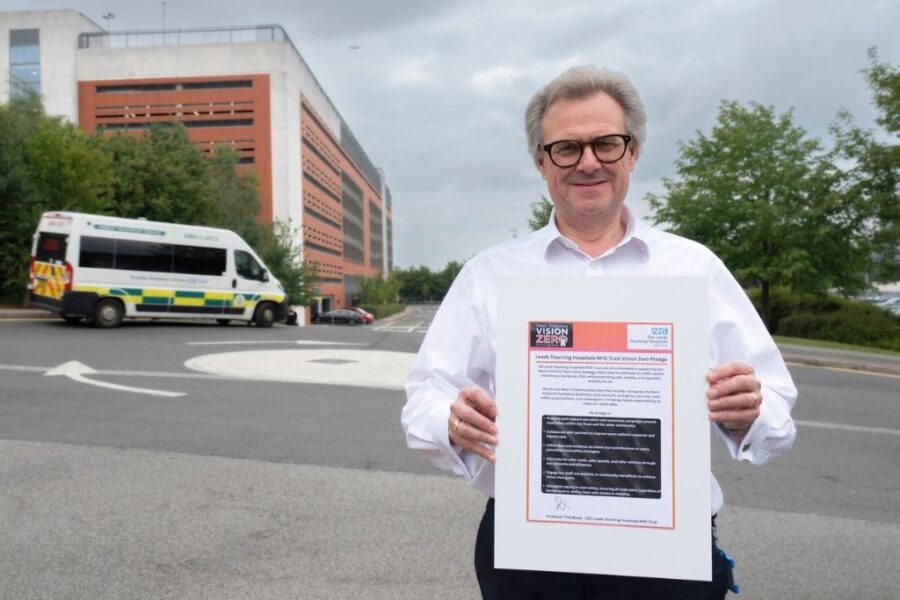 A man in a white shirt stood holding a placard with a series of pledges to do with the Vision Zero strategy. He is stood in front of a roundabout with an ambulance in the background