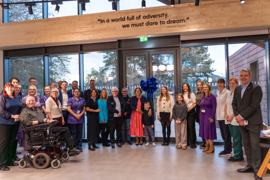 group of people standing in front of glass doors with a large blue ribbon behind them. There is a beam above with the inscription "In a world full of adversity, we must dare to dream." - Rob Burrow CBE.