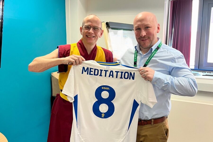 A man in yellow and burgundy robes stood next to a man in a blue shirt holding a football shirt with the number 8 and 'meditation' printed on it.