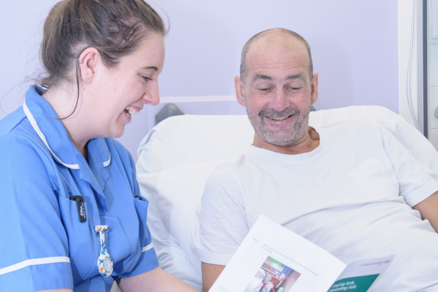 A woman in a blue clinical uniform sat reading a leaflet to a man in a white t shirt lying on a bed.