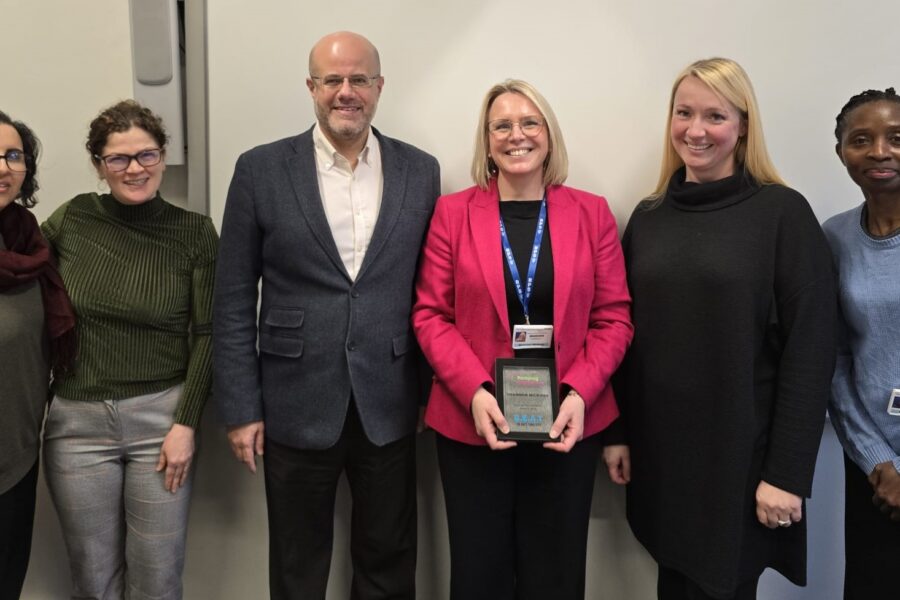 A group of seven people stand side by side indoors in front of a light‑coloured wall. The person in the center (Shannon Nixon) is holding her award plaque with both hands.