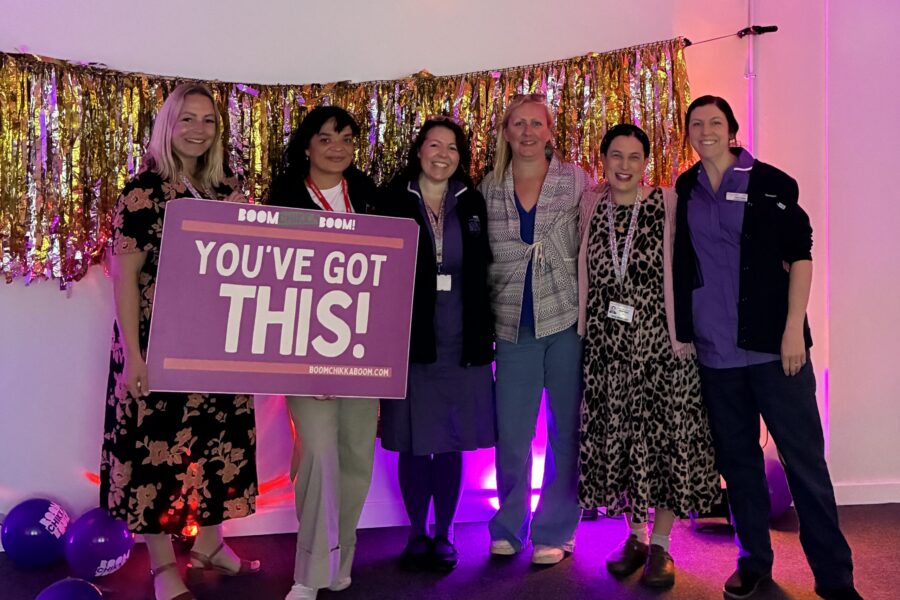 Midwives and members of the Baby Bank team stood in a line holding a purple sign against a glittery backdrop and purple disco lights.