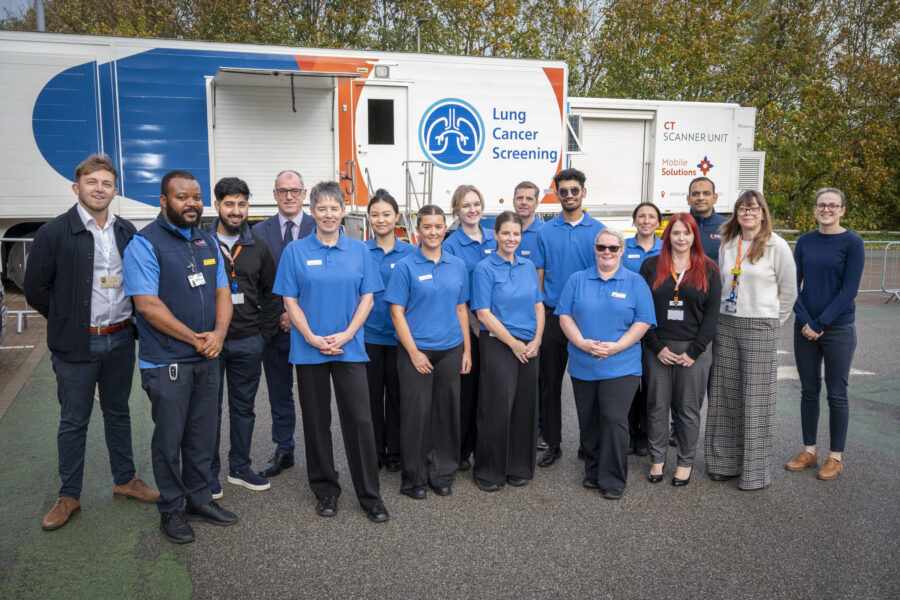 A group of people, some in blue clinical uniforms stood outside a white mobile scanning trailer in a car park.