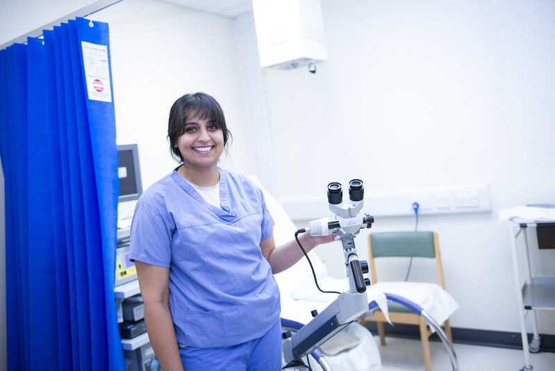 A consultant in blue scrubs stood next to a gynaecology microscope in a clinical room with a dark blue curtain next to the couch.