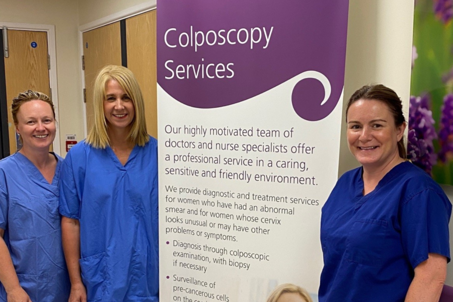 Three nurses in blue scrubs stood next to a banner that says Colposcopy services