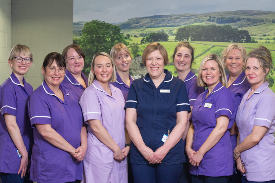 Photo of research team wearing purple, lilac and navy uniforms. Group of 11 midwives and research assistants stood against a background of Yorkshire scenery.